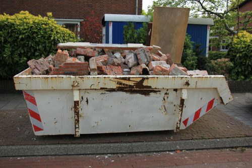 A large, white metal skip positioned on a paved street in an urban area, filled with a mixture of red bricks, concrete fragments, and broken building materials. The surface of the skip shows signs of rust and weathering, with some dark streaks and stains. The surrounding environment includes a driveway with a brick border, a garden with green shrubbery on the left, and a blue shed or garage partially visible in the background. The scene appears to be during daylight hours, with natural light illuminating the debris and the environment, suggesting a recent rubbish removal or renovation activity. The image is relevant to commercial waste removal services provided by companies like Commercial Waste Removal Shadwell, operating within the postcode area around Shadwell, London, placeholder for local context in waste disposal operations.