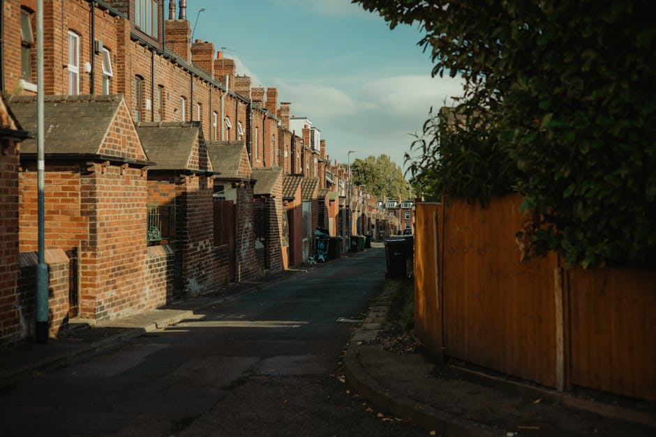 A narrow residential alleyway lined with traditional red-brick terraced houses on the left, each with small front walls, sloped tiled roofs, and chimneys, casting long shadows on the tarmac pavement.