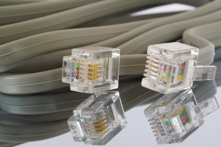 A close-up view of four clear plastic Ethernet connectors with gold-plated contacts, placed on a reflective surface. Behind them, a coiled bundle of grey Ethernet cable with a smooth, flexible texture